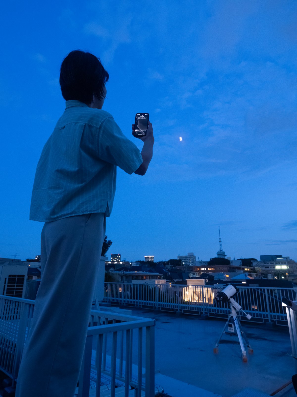 夕暮れのベランダでスマートフォンをかざし、月をステラナビゲータ・モバイルで確認している様子。かたわらには望遠鏡も置かれている